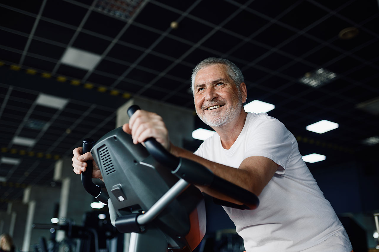 Elderly man poses on exercise bike, gym interior on background. Sportive grandpa on fitness training in sport center. Healty lifestyle, health care, old sportsman
